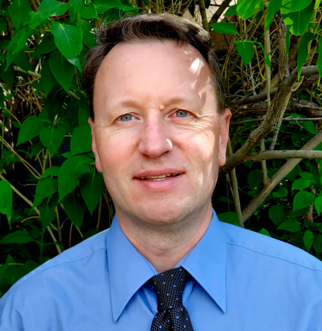 A head shot of Michael in a blue shirt and tie against green brush