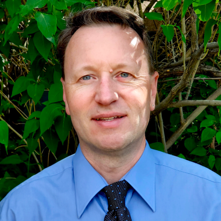 A head shot of Michael in a blue shirt and tie against green brush